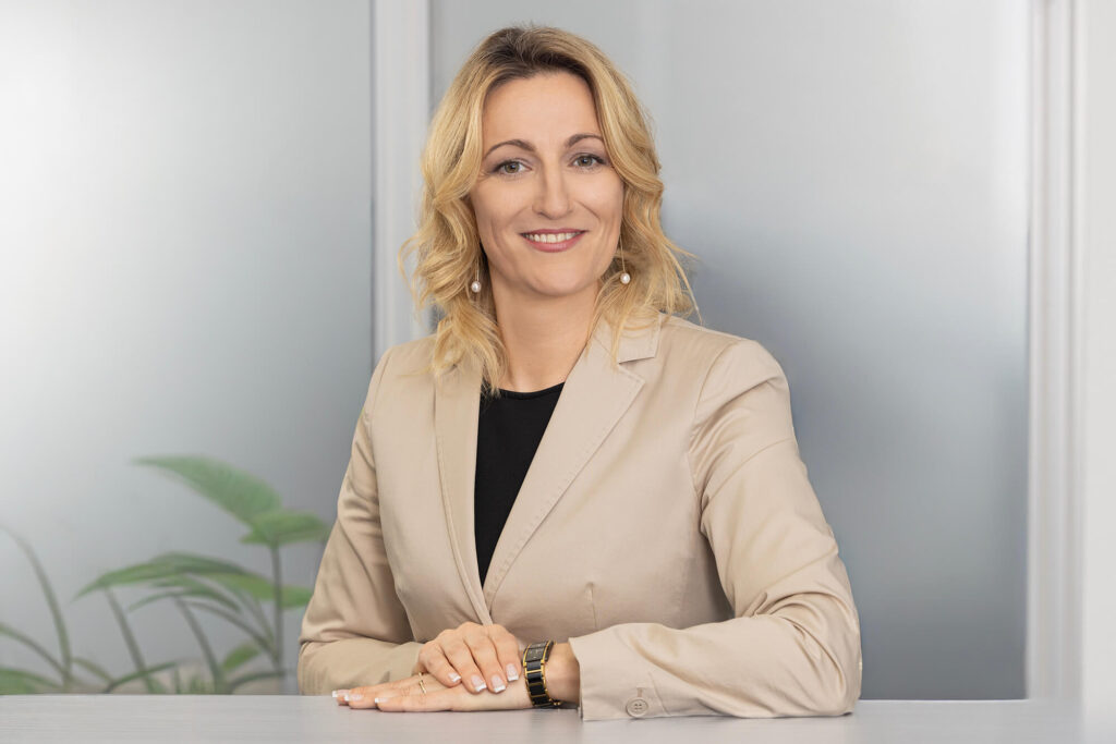 Portrait Elena Nestmeyer im beigen Blazer auf die Empfangstheke gelehnt, freundlich lächelnd